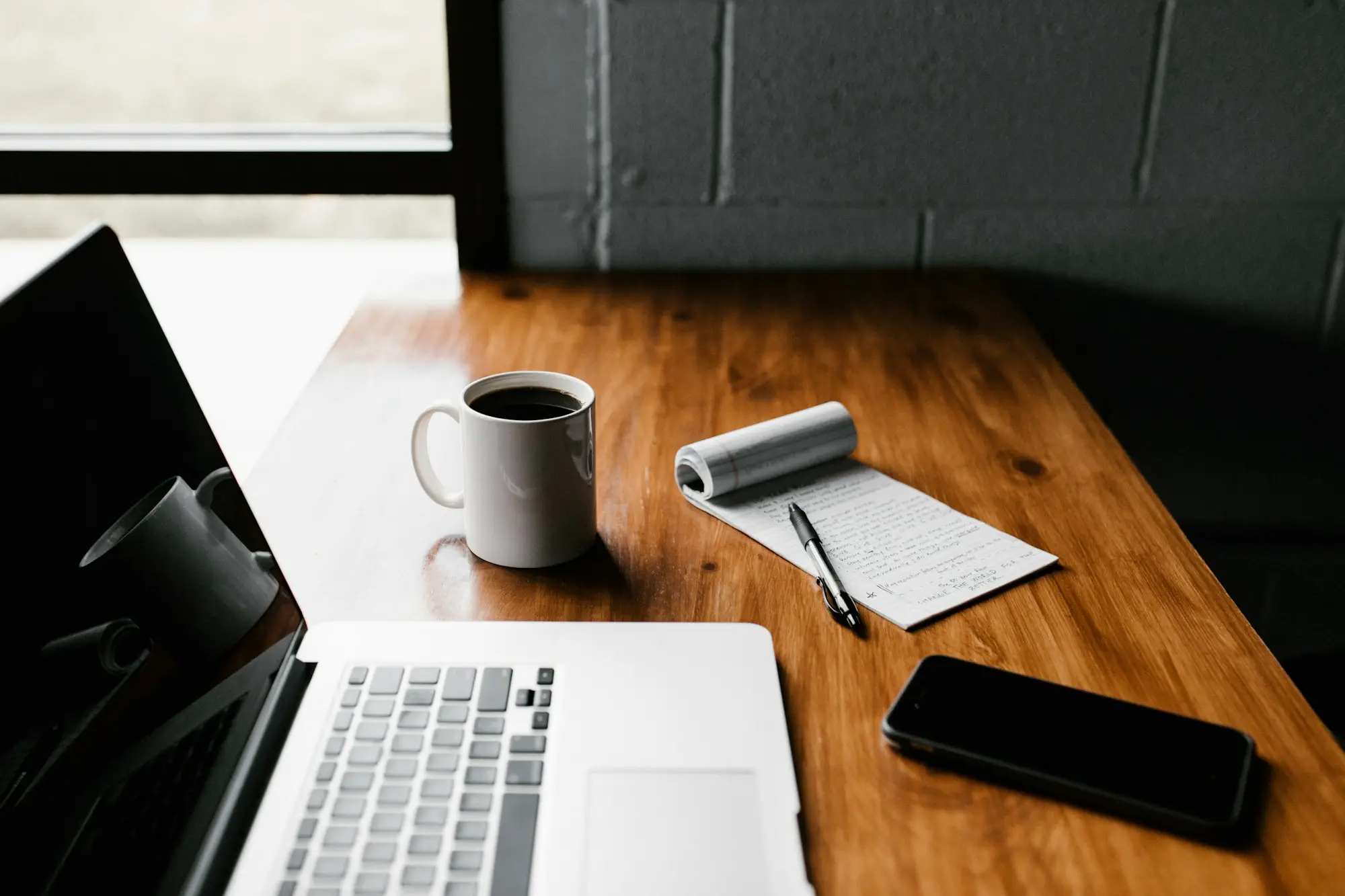 A laptop, notepad, pen and coffee cup arranged on a wooden desk ready for writing